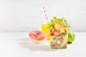 Healthy take away food set with fresh orange juice, tropical shrimp salad with lettuce, red bell pepper, croutons, green apple, pink donut in plastic pack in sunny white interior with marble wall.