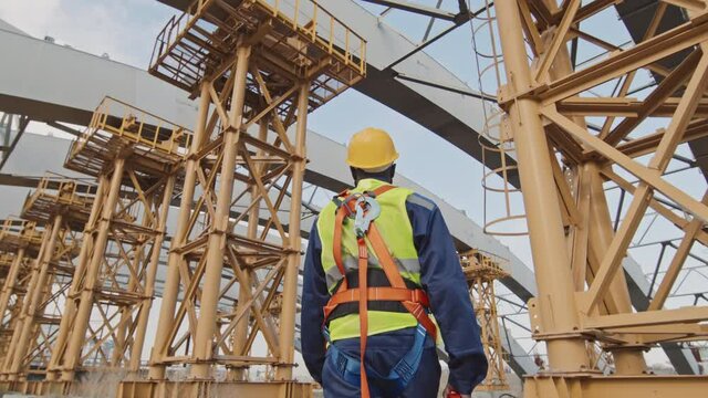 Low angle shot of unrecognizable African-American male worker in hard hat and safety belt with harness walking at construction site with unfinished building with scaffolding