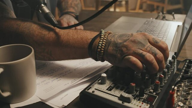 Tracking close up shot of unrecognizable male podcast host with tattoos using audio mixer while recording his show