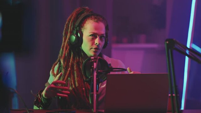 Tracking shot of young woman with dreadlocks sitting at table in studio lit by neon string lights and talking into microphone during recording of podcast