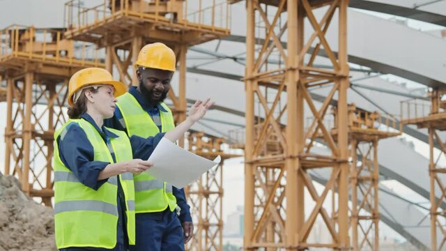 Tracking shot of Caucasian female and African-American male construction workers in safety vests and hard hats walking before unfinished building with scaffolding and discussing blueprint