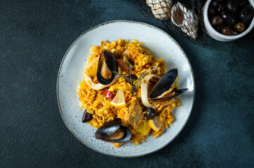 Classic dish of Spain, seafood paella in plate on blue background top view. Spanish paella with shrimps, clamps, mussels and fresh lemon. Spanish food. Comfort food. Rice with seafood.