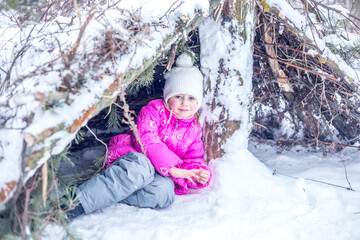 Little girl plays in a hut from coniferous branches in the winter forest