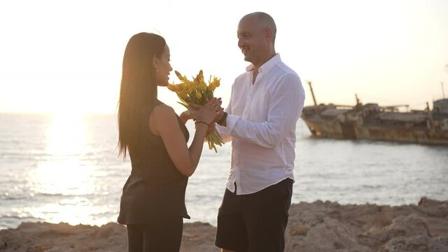 Happy smiling loving man surprising woman with bouquet of flowers standing in sunbeam on Mediterranean sea coast. Side view of Caucasian husband and pregnant wife enjoying sunset outdoors