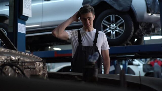 Front view of exhausted mechanic male in blue uniform rubbing head with hands and sighing. Handsome tired Caucasian young service man standing near car with open bonnet in auto repair shop indoors.