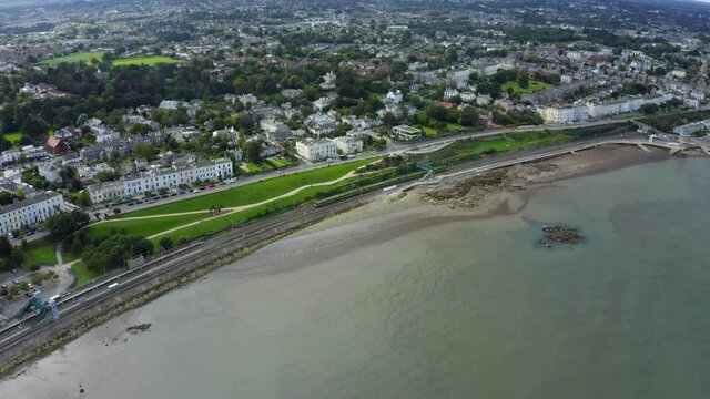 Salthill and Monkstown Station, Dublin, Ireland, September 2021. Drone tracks parallel to the coastline as a southbound Dart train arrives at the station.