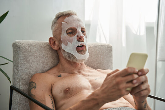 Man with facial mask sitting at the armchair and looking at the smartphone - Powered by Adobe