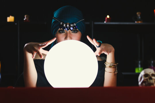 Mysterious magnificent beautiful woman fortune teller in black dress reading future on luminous crystal ball, dark witch casting a spell with magical ball.