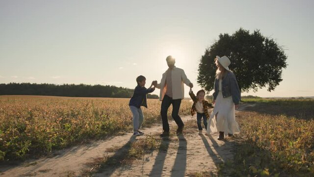 Joyful family of married adult caucasian couple holding hands with cute kids, walking in natural countryside field outside. Adorable scene. Relationships.