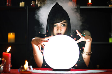 Dark witch in black hood casting spell on luminous magical ball with smoke light background,...