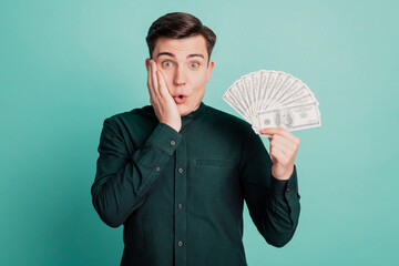 Portrait of surprised funny millionaire guy hold fan cash palm cheek wow reaction on turquoise...