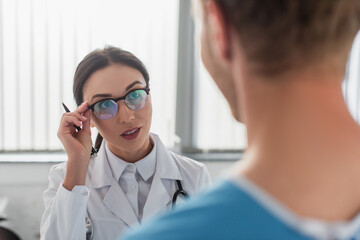 brunette doctor with pen adjusting glasses while looking at blurred patient in hospital