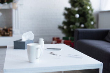 table with paper napkins, pills container, electronic thermometer and smartphone near blurred christmas tree