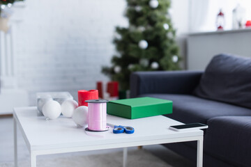 table with gift box, bobbin with decorative ribbon, baubles and scissors in living room with blurred christmas tree