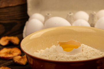 A broken white chicken egg in a bowl with wheat flour, dried apple chips and several whole eggs in a tray made of white cardboard, close-up selective focus.