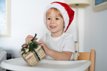 Joyful baby in santa hat holding a gift box at home