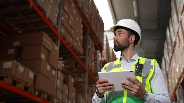 Close-up of handsome worker in hard hat and vest using tablet while checking products stock in warehouse. Bearded storehouse employee doing stocktaking while walking among shelves with cardboard boxes