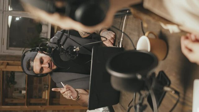 Handheld vertical shot of man and woman in headphones sitting at table at home and having discussion while recording their podcast
