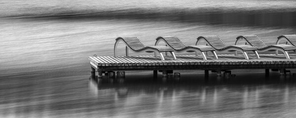 Row of empty deck chairs on dock at lake. Wooden bridge with sun-beds and observation space. Black and white photograph of relaxation, vacation and rest in nature  concept