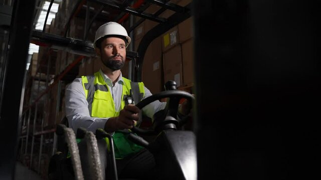 Portrait of confident bearded man wearing hard hat and vest riding loader between racks with cardboard boxes. Portrait of serious forklift driver during work in warehouse