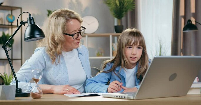 Nice positive confident caring blond mother in glasses helping her smart daughter with homework, sitting at workplace and reading tasks on laptop screen,close up