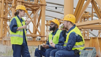 Medium shot of cheerful female and male workers in safety vests and hard hats eating lunch by scaffolding at construction site and chatting