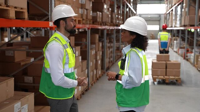 Two logistics managers, mixed race female and caucasian male, wearing hard hats and vests meeting in storehouse to discuss work issues. Pair of warehouse employees communicating at meeting