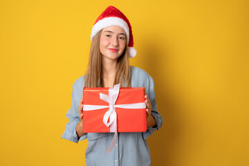 Young smiling woman in Santa hat holding christmas gift box isolated over yellow background.