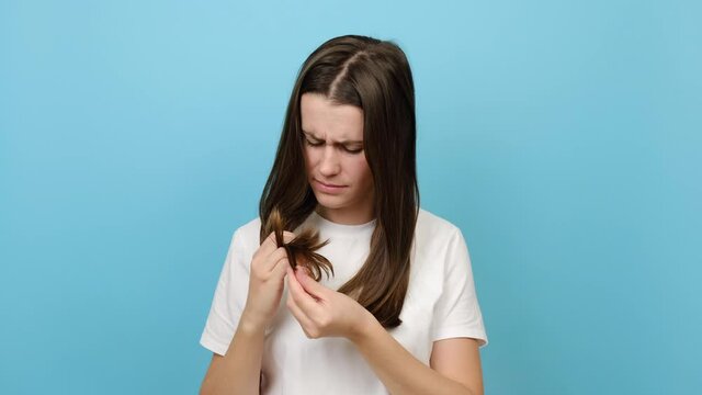 Depressed young woman brushing looking at split ends, isolated over blue studio background. Worried sad girl feels upset about brittle damaged dry hair loss concept. Female hormone problems concept