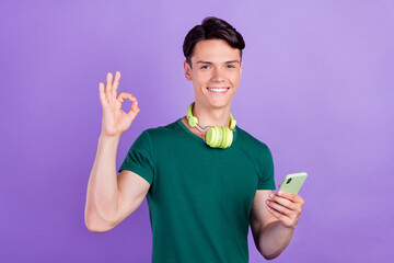 Photo of brunette positive young man hold phone okay sign headphones isolated on violet color background