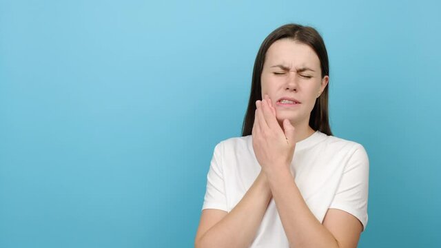 Unhappy sad young caucasian woman suffering from acute tooth pain, touching her sore jaw and frowning, isolated over blue studio background wall with copy space. Oral hygiene and toothcare concept