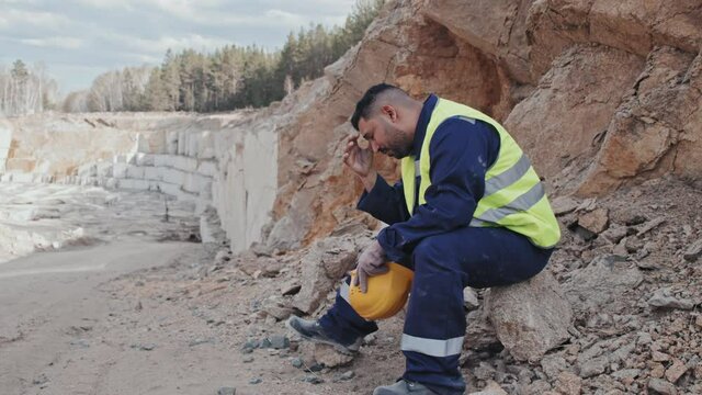 Handheld tracking of tired Arab male worker in uniform and hard hat sitting on stone in granite quarry and resting