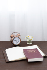 Bible and notebook on the table with an alarm clock by the window. Vertical format