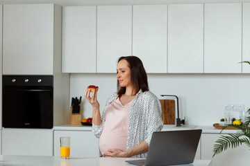 Brown haired young pregnant woman holds delicious cupcake standing near table with orange juice and laptop on top in kitchen