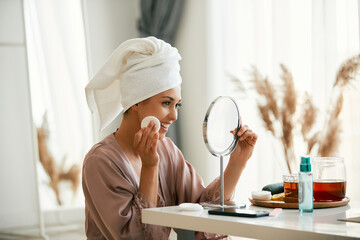 Happy woman looks herself in mirror and cleans her face skin with cotton pad.
