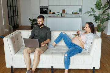 Young pregnant woman lies on white sofa holding plate with food near sitting husband with laptop against spacious light room