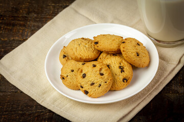 Pile of Chocolate chip cookie on white plate and glass of milk on rustic wooden background.