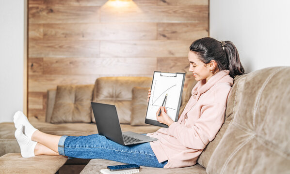 Girl at home sitting on the sofa with laptop and showing graph on clipboard at online meeting.