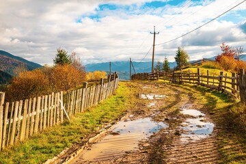 A path along the ridge of a hill with a large puddle and an old fence against the backdrop of a colorful forest