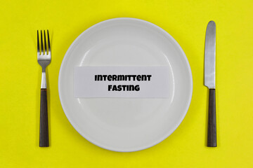 Table setting, empty white plate with fork and knife on yellow background, with one of the principles of healthy living - Intermittent fasting