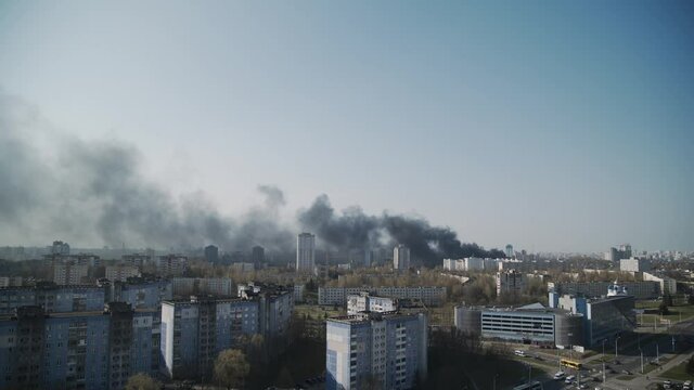 Fire in big metropolis in spring, panoramic city view with blue sky. Black smoke rises above rooftops of high-rise buildings, covers town. Air pollution, environmental disaster. Cars driving on road.