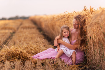 Mother and little daughter have fun sitting by the golden wheat field. Young woman tickles the girl