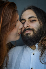 Portrait of a young couple where a long red haired woman kisses the cheek of the man of the bearded man with long hair