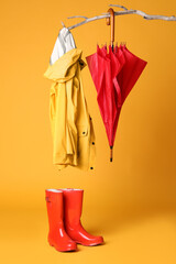 Closed red umbrella, stylish raincoat and rubber boots hanging on branch against yellow background