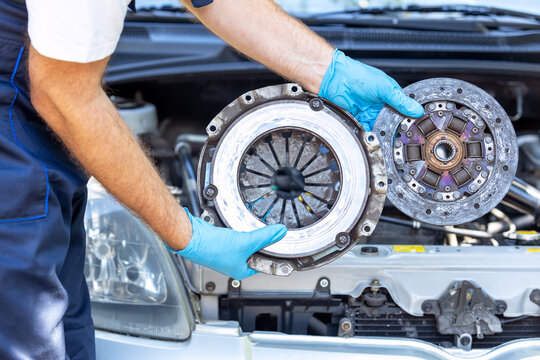 Automotive technician holding used car pressure plate and clutch disc in front of the vehicle engine