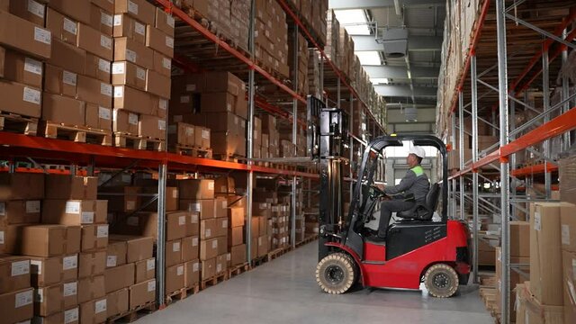 Storehouse worker wearing hard hat taking pallet of cardboard boxes from shelf and riding forklift truck between rows. Male driver skillfully operating forklift in factory storage