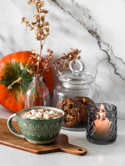 Hot chocolate with marshmallows and pumpkin pie spices on the table, a jar of cookies, a lit candle and a pumpkin. Cozy kitchen interior