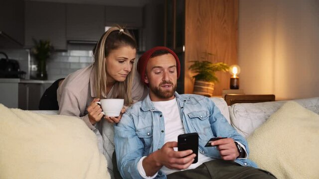 happy caucasian couple sitting on couch making online credit card payment on cellular device 