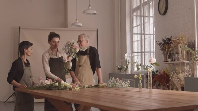 Wide stab shot of creative young female florist teaching floral masterclass in cozy studio to kind adult couple