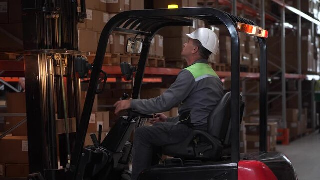 Focused man wearing hard hat working on forklift in factory storage. Experienced operator of warehouse loader during work, riding, lifting, placing goods on racks filled with cardboard boxes